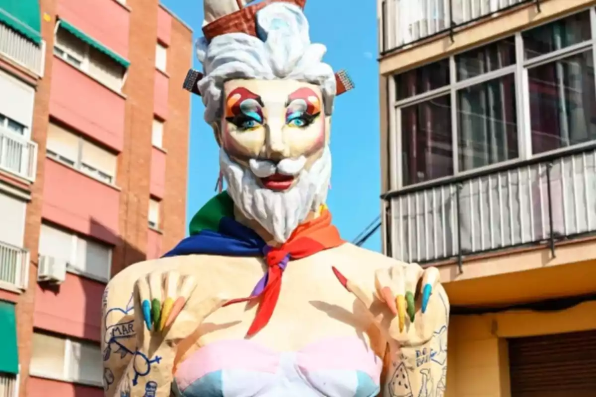 Figura de cartró amb barba blanca, maquillatge colorit, ungles pintades de colors i mocador arc de Sant Martí al coll, situada en un carrer entre edificis.