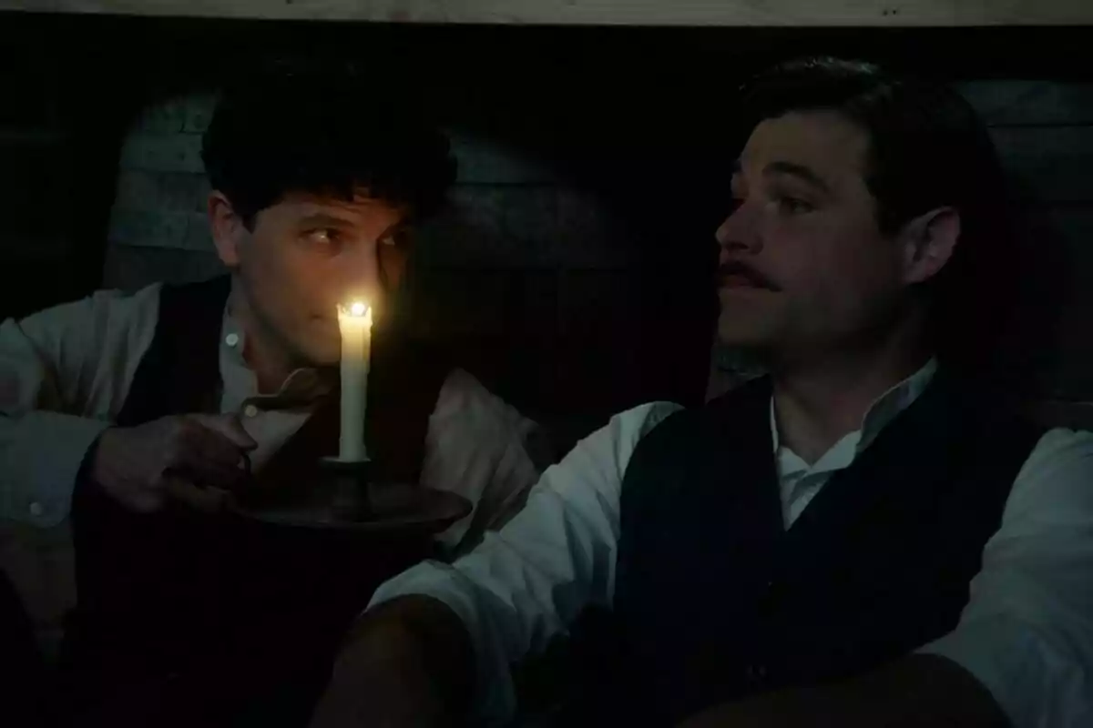Dos homes asseguts en un lloc fosc, Álvaro Quintana subjectant una espelma mentre mira Arturo Sancho a La Promesa Dos homes asseguts en un lloc fosc, Álvaro Quintana subjectant una espelma mentre mira Arturo Sancho a La Promesa