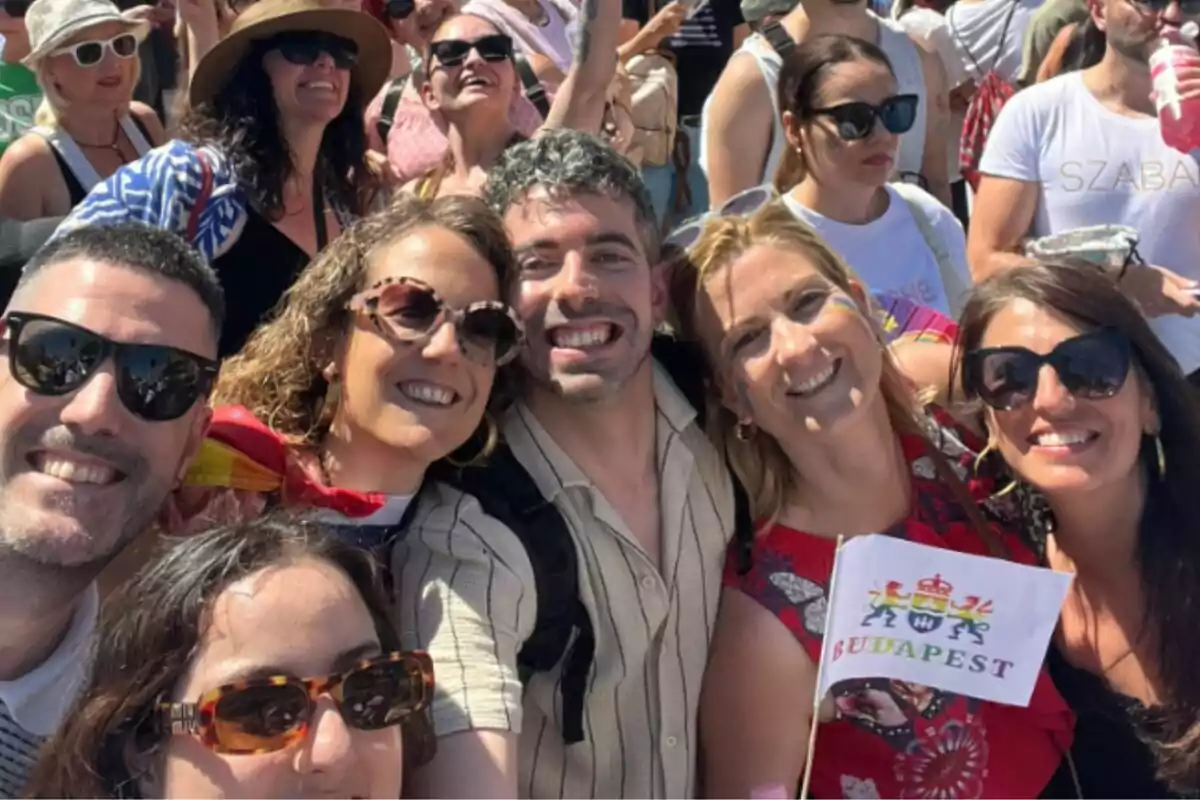 Grup de persones somrients amb ulleres de sol gaudint d’un esdeveniment a l’aire lliure, una d’elles sosté una bandera que diu Budapest
