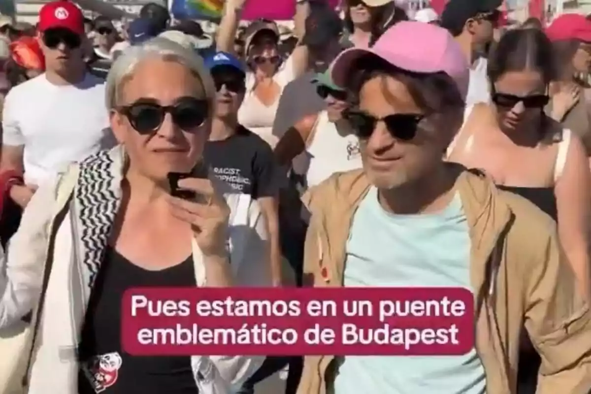 Dues persones amb ulleres de sol i roba informal són al mig d’una multitud durant un esdeveniment a l’aire lliure, amb un text que diu Dues persones amb ulleres de sol i roba informal són al mig d’una multitud durant un esdeveniment a l’aire lliure, amb un text que diu
