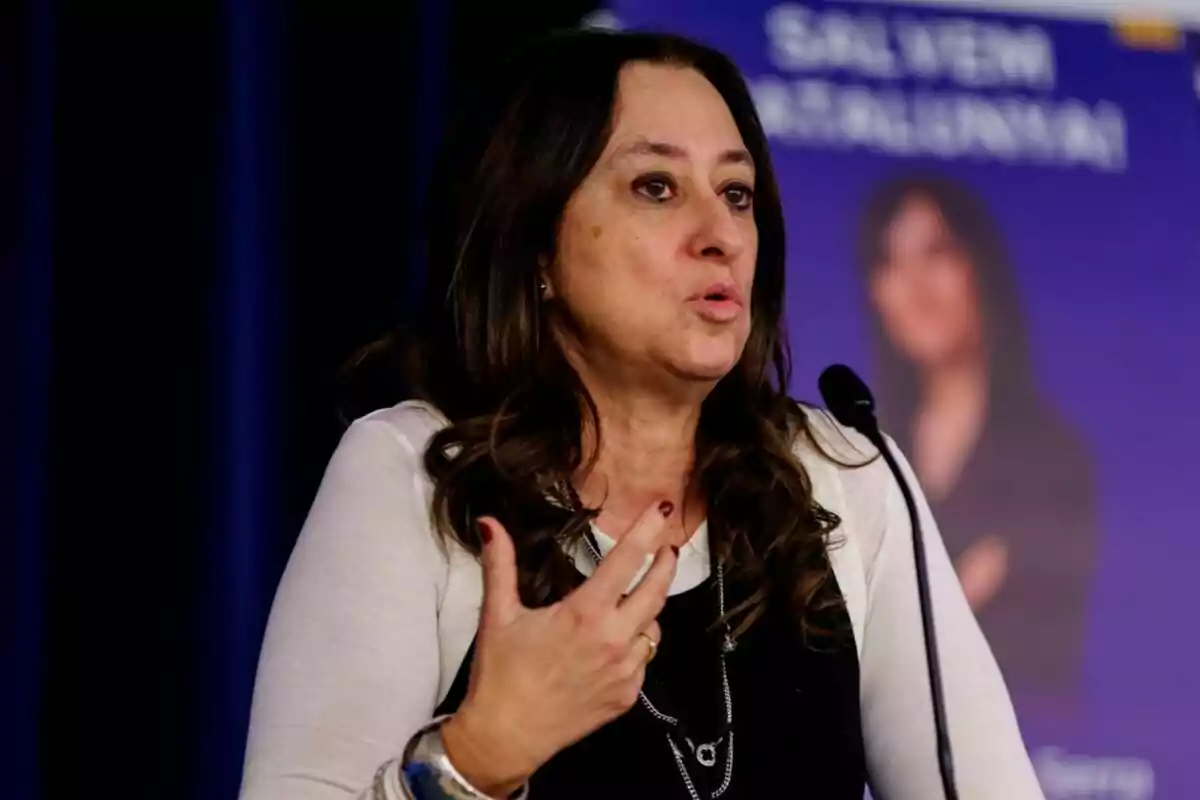 Woman with long dark hair speaking in front of a microphone with a blue background and a blurred sign behind her