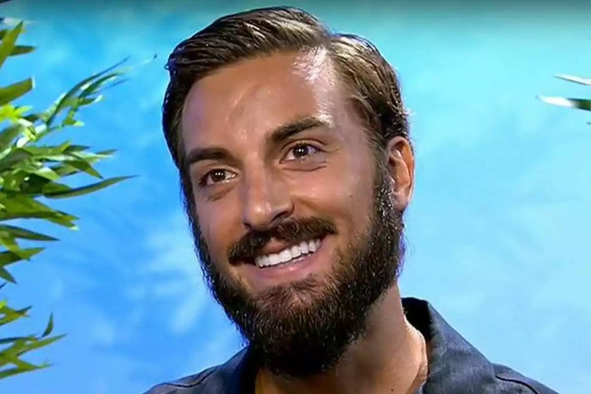 Borja González, un jove amb barba i cabell curt, somriu davant d’un fons blau amb plantes als costats a Supervivientes.