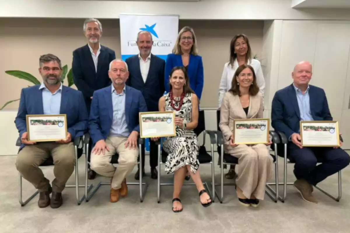 Grup de persones posant en una sala d’oficina, algunes assegudes i d’altres dretes, diverses sostenen diplomes emmarcats i al fons hi ha un cartell de la Fundació La Caixa. Grup de persones posant en una sala d’oficina, algunes assegudes i d’altres dretes, diverses sostenen diplomes emmarcats i al fons hi ha un cartell de la Fundació La Caixa.