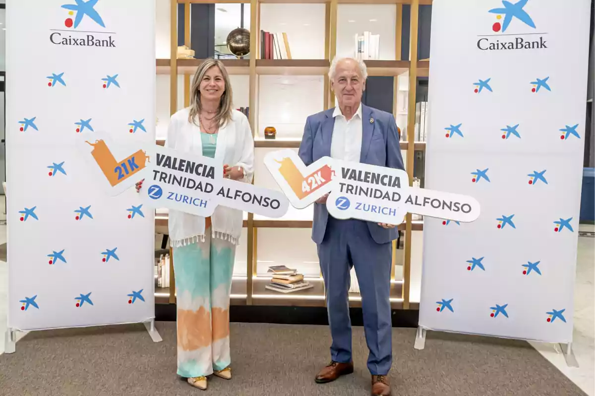 Dues persones posen somrients en un esdeveniment de CaixaBank sostenint cartells promocionals de les curses 21K i 42K València Trinidad Alfonso Zurich, amb un fons d'estanteria i panells amb el logotip de l'entitat bancària. Dues persones posen somrients en un esdeveniment de CaixaBank sostenint cartells promocionals de les curses 21K i 42K València Trinidad Alfonso Zurich, amb un fons d'estanteria i panells amb el logotip de l'entitat bancària.