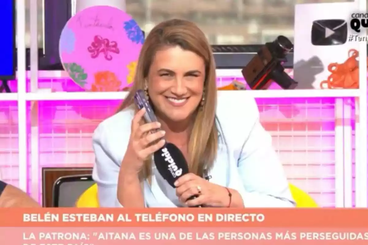 Carlota Corredera somrient a 'Tentacles' sostenint un telèfon mòbil i un micròfon mentre està asseguda en un plató colorit amb objectes decoratius al fons Carlota Corredera somrient a 'Tentacles' sostenint un telèfon mòbil i un micròfon mentre està asseguda en un plató colorit amb objectes decoratius al fons