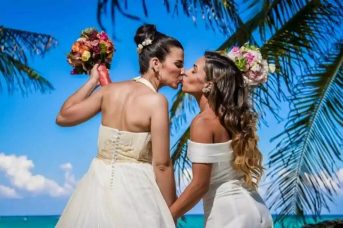 Dues dones vestides de núvia es fan un petó a la platja sota palmeres i un cel blau, totes dues sostenen rams de flors colorides a Casats a primera vista. Dues dones vestides de núvia es fan un petó a la platja sota palmeres i un cel blau, totes dues sostenen rams de flors colorides a Casats a primera vista.