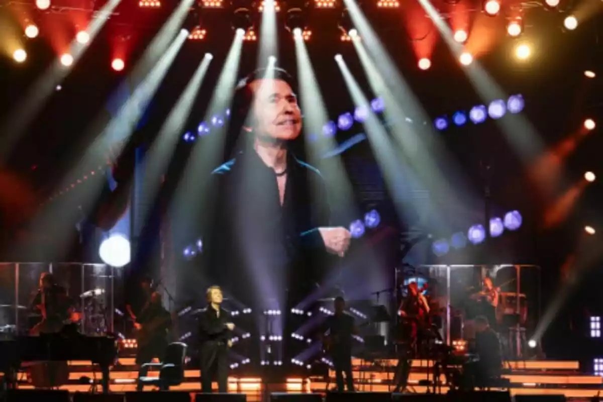 Raphael performs on a stage lit with colorful lights, and a large screen in the background displays his image while the band plays behind him. Raphael performs on a stage lit with colorful lights, and a large screen in the background displays his image while the band plays behind him.