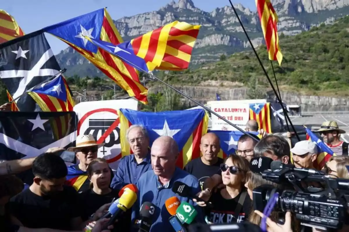 Un grup de persones es manifesta a l’aire lliure amb banderes estelades i micròfons de diversos mitjans de comunicació, amb muntanyes al fons. Un grup de persones es manifesta a l’aire lliure amb banderes estelades i micròfons de diversos mitjans de comunicació, amb muntanyes al fons.