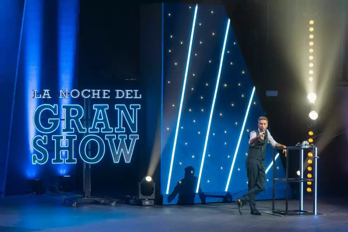 Dani Martínez dret en un escenari il·luminat amb llums blaves i un rètol que diu La Nit del Gran Show Dani Martínez dret en un escenari il·luminat amb llums blaves i un rètol que diu La Nit del Gran Show