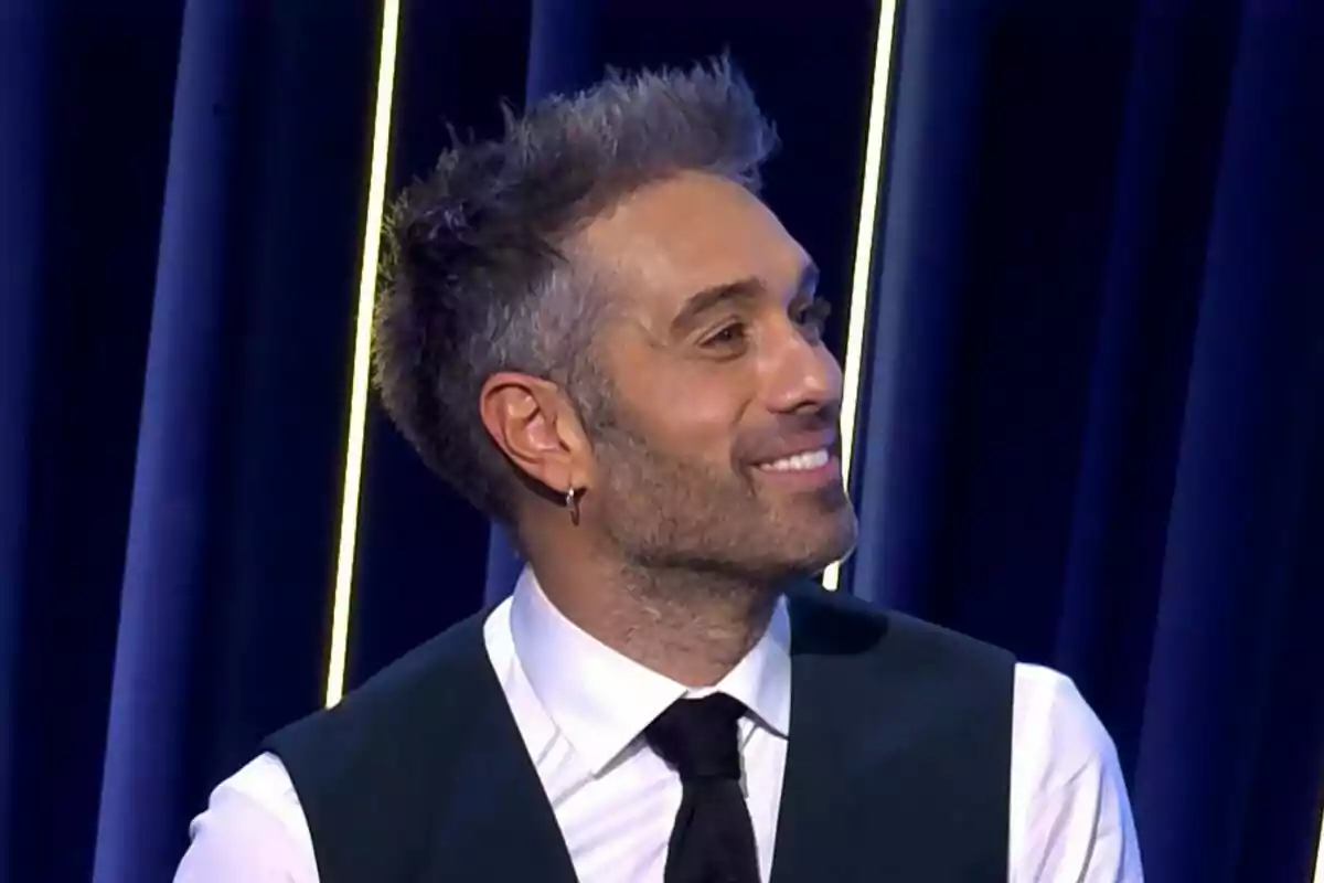 Dani Martínez de cabell curt i barba lleugera somrient, vestit amb camisa blanca, armilla fosca i corbata negra, davant d’un fons blau amb línies verticals il·luminades a El Gran Show.