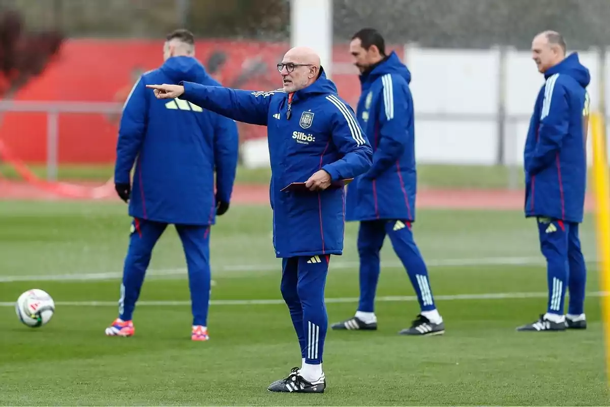 Entrenador de futbol donant indicacions en un camp d’entrenament acompanyat d’altres membres del cos tècnic, tots vestits amb roba esportiva blava Entrenador de futbol donant indicacions en un camp d’entrenament acompanyat d’altres membres del cos tècnic, tots vestits amb roba esportiva blava