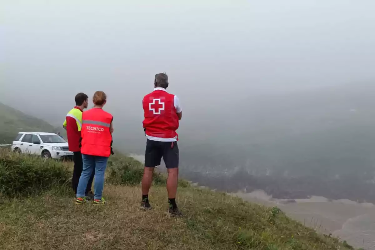 Tres persones amb armilles d’emergència observen un penya-segat cobert de boira al costat d’un vehicle blanc estacionat a l’herba.