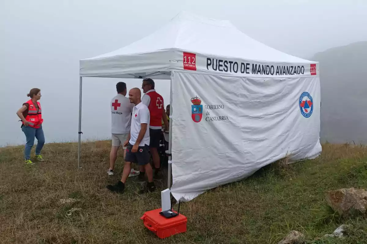 Diverses persones amb armilles i samarretes de la Creu Roja estan reunides al costat d’una carpa blanca de punt de comandament avançat en una zona de camp amb boira. Diverses persones amb armilles i samarretes de la Creu Roja estan reunides al costat d’una carpa blanca de punt de comandament avançat en una zona de camp amb boira.