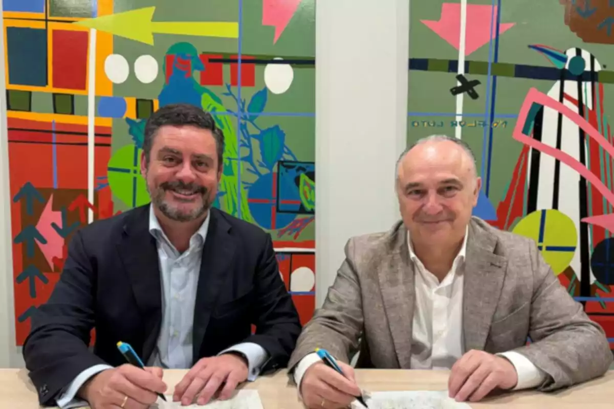 Dos homes somrients asseguts en una taula signant documents amb bolígrafs davant d’un mural abstracte i colorit Dos homes somrients asseguts en una taula signant documents amb bolígrafs davant d’un mural abstracte i colorit
