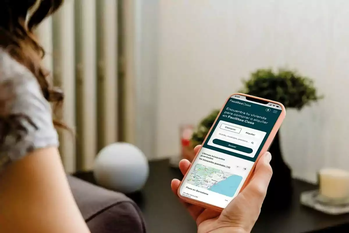 Persona sostenint un telèfon mòbil amb una aplicació de cerca d’habitatges oberta a la pantalla, asseguda en una habitació amb decoració minimalista.