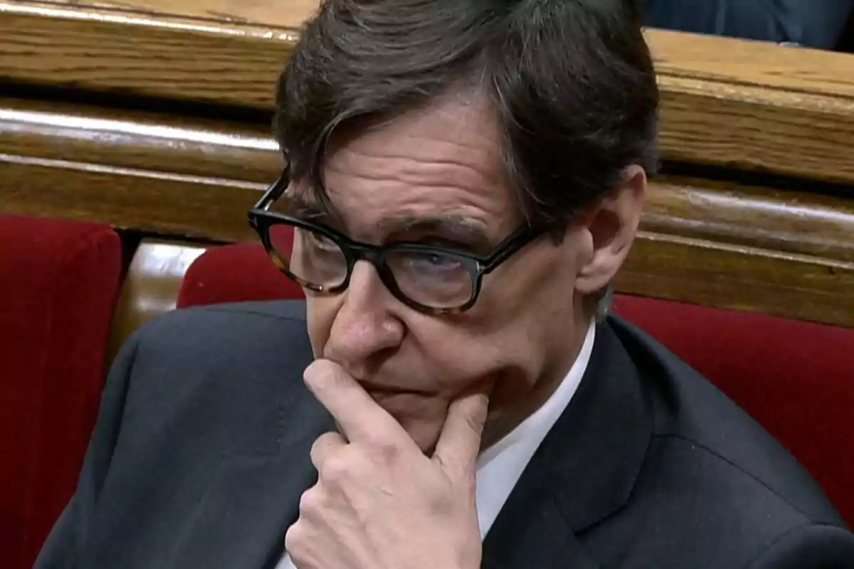 Home amb ulleres i vestit fosc assegut en un banc vermell amb expressió pensativa i la mà al rostre