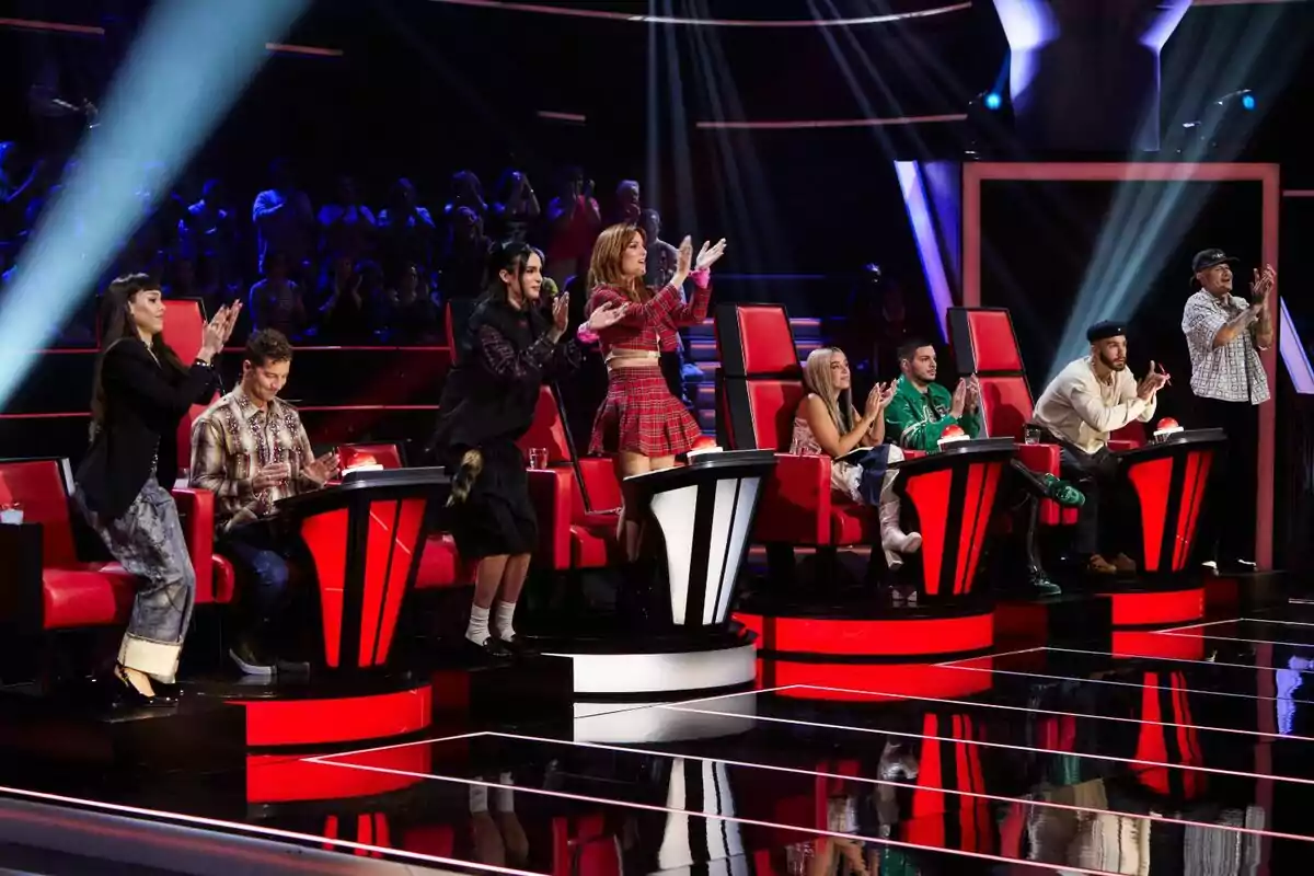 Diversos jutges asseguts i drets aplaudeixen en un plató de televisió amb cadires vermelles i llums brillants mentre el públic observa al fons de La Voz Kids. Diversos jutges asseguts i drets aplaudeixen en un plató de televisió amb cadires vermelles i llums brillants mentre el públic observa al fons de La Voz Kids.