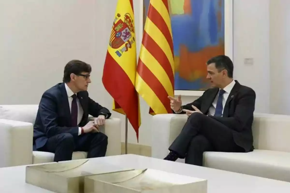Dos homes amb vestit conversen asseguts en butaques blanques amb les banderes d’Espanya i de Catalunya darrere seu.