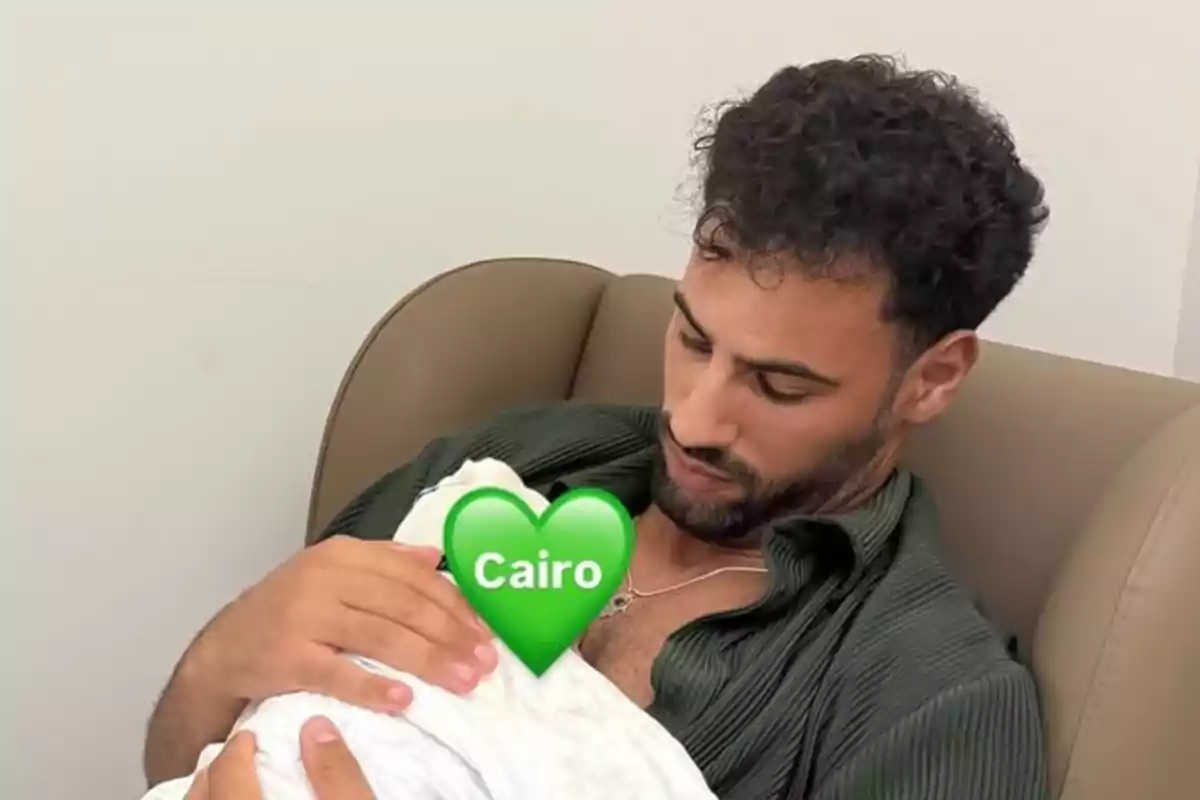 Man sitting in an armchair holding a baby wrapped in a white blanket with a green heart emoji that says Cairo covering the baby's face Man sitting in an armchair holding a baby wrapped in a white blanket with a green heart emoji that says Cairo covering the baby's face