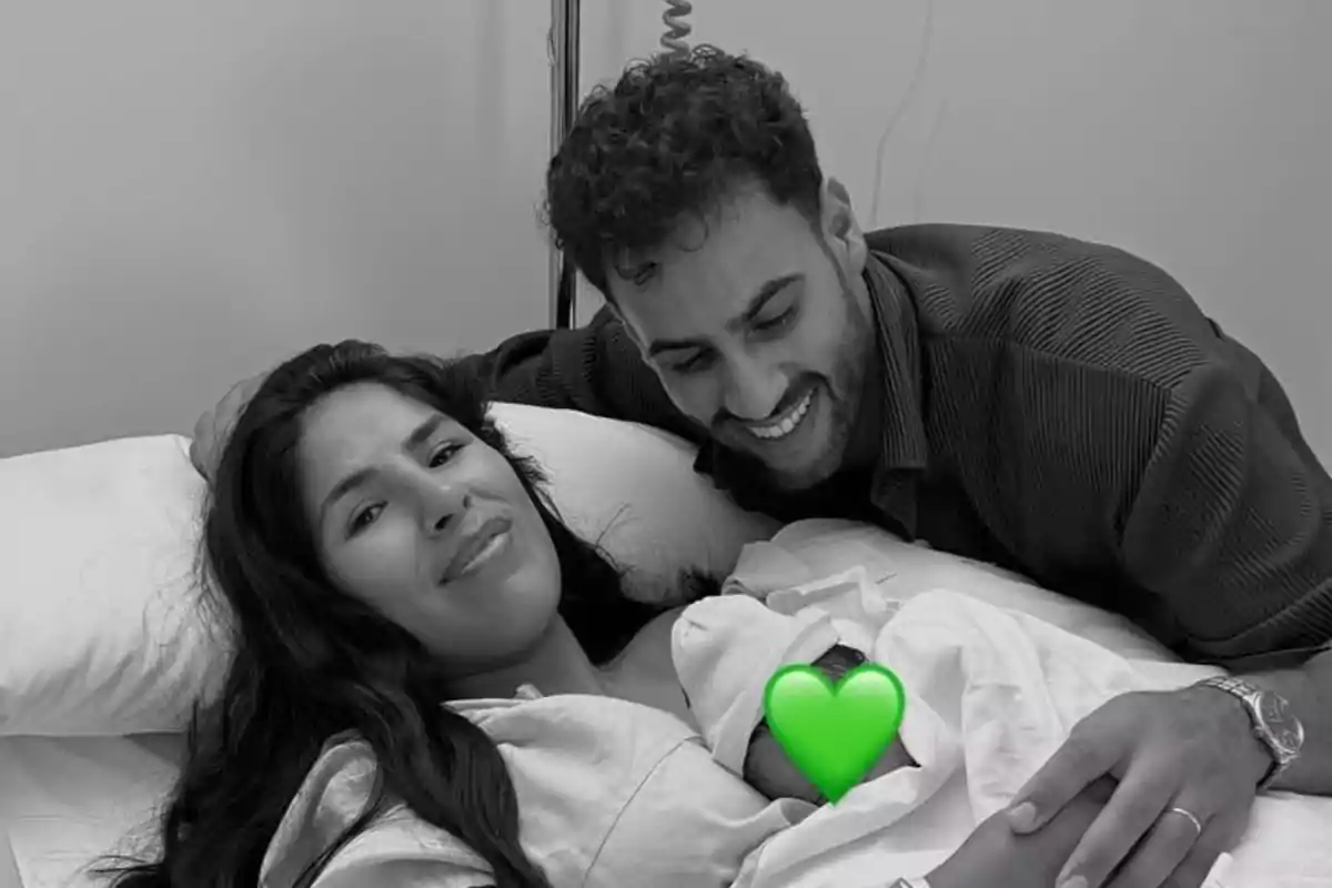 A couple smiles while holding their newborn baby on a hospital bed, with a green heart covering the baby. A couple smiles while holding their newborn baby on a hospital bed, with a green heart covering the baby.