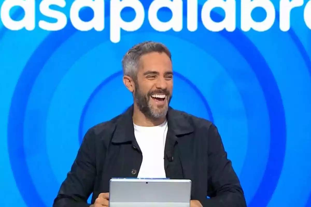 Short-haired man with a beard smiling while holding a tablet in front of a blue background with white letters Short-haired man with a beard smiling while holding a tablet in front of a blue background with white letters