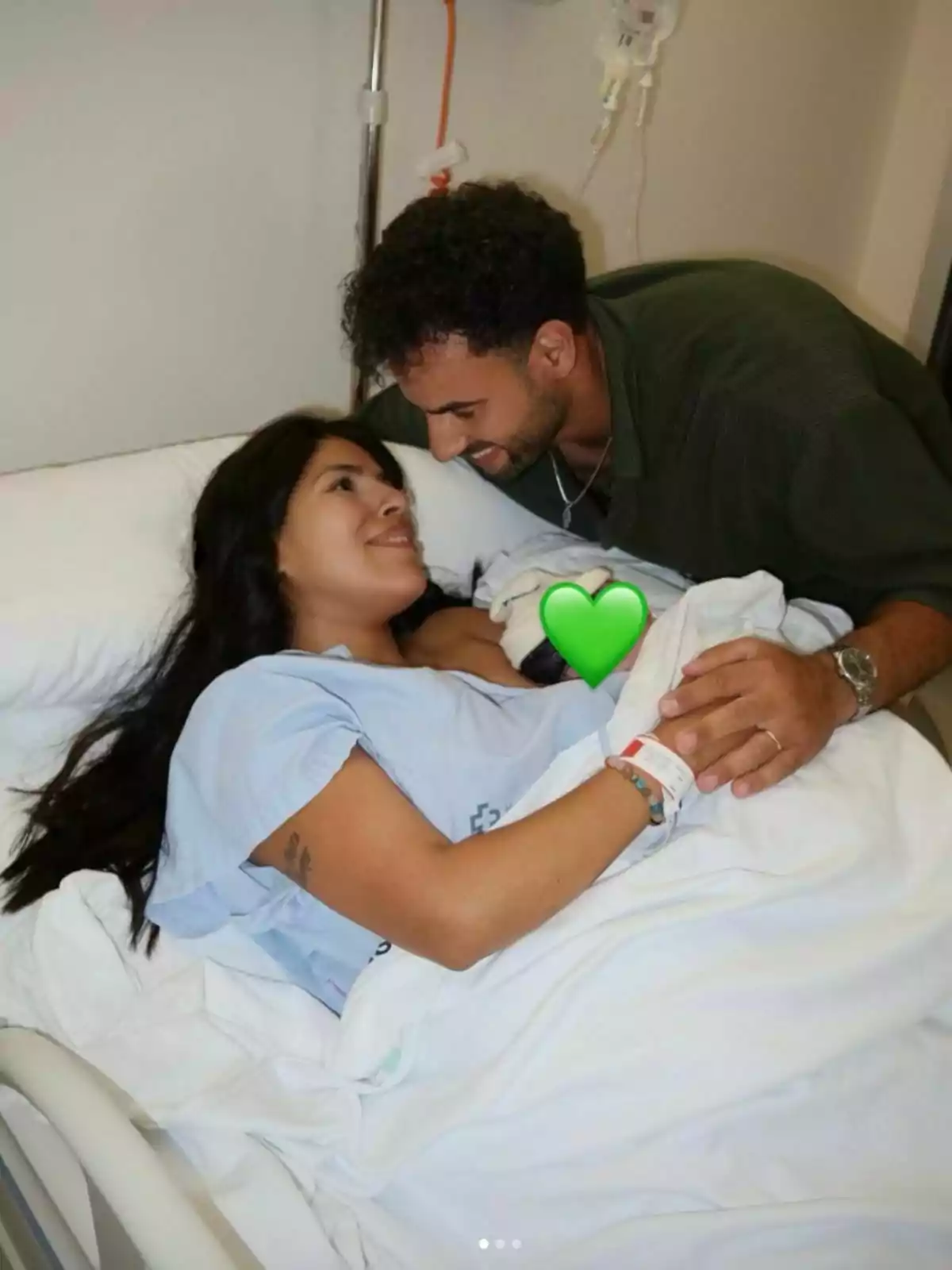 Isa Pantoja lying on a hospital bed smiles while Asraf Beno looks at her tenderly and they hold hands, covered by a white sheet. Isa Pantoja lying on a hospital bed smiles while Asraf Beno looks at her tenderly and they hold hands, covered by a white sheet.