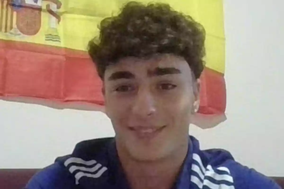 Young man in a blue t-jersey smiles in front of a Spanish flag on the wall.