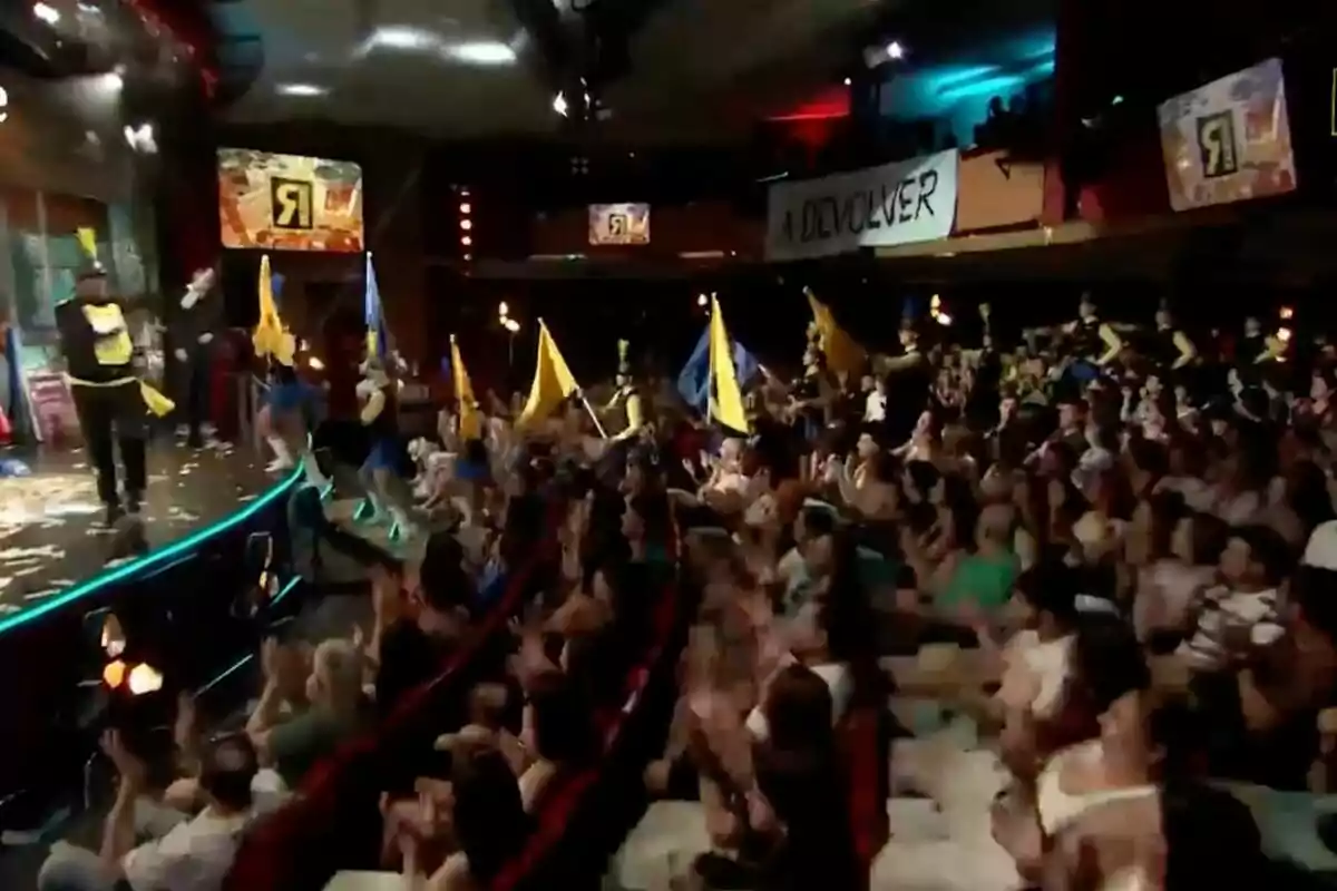 Un teatre ple de persones aplaudint mentre un grup d’animadors amb banderes grogues i blaves es presenta a l’escenari a ‘La Revuelta’. Un teatre ple de persones aplaudint mentre un grup d’animadors amb banderes grogues i blaves es presenta a l’escenari a ‘La Revuelta’.
