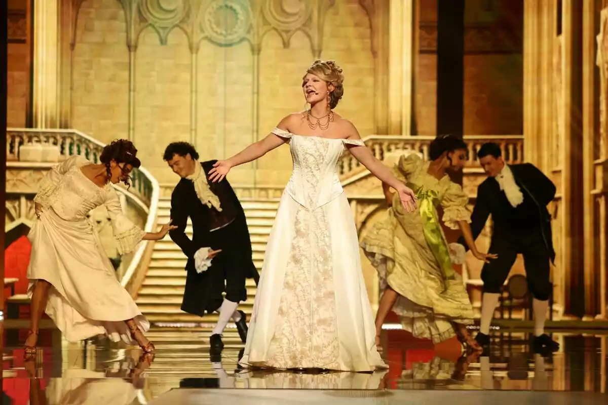 Laura Escanes amb vestit elegant canta al centre d’un escenari mentre quatre ballarins amb vestits d’època l’envolten en un ambient que simula un palau antic a Tu Cara Me Suena. Laura Escanes amb vestit elegant canta al centre d’un escenari mentre quatre ballarins amb vestits d’època l’envolten en un ambient que simula un palau antic a Tu Cara Me Suena.