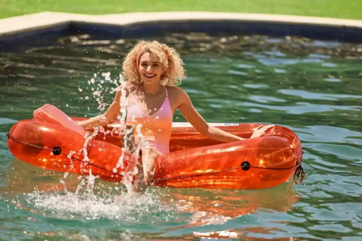 Dona somrient amb banyador rosa asseguda en un inflable taronja en una piscina mentre esquitxa aigua Dona somrient amb banyador rosa asseguda en un inflable taronja en una piscina mentre esquitxa aigua
