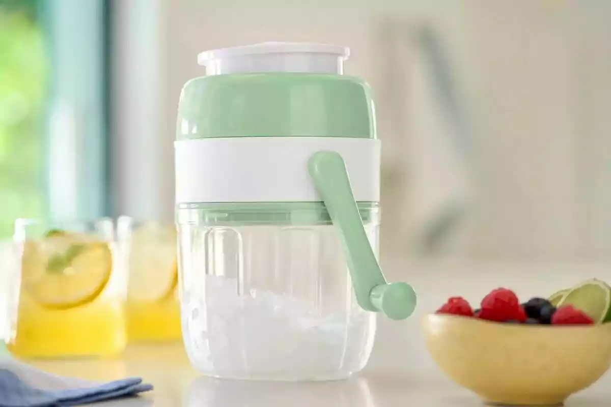 Green and white manual ice crusher on a table next to drinks and a bowl of fruit Green and white manual ice crusher on a table next to drinks and a bowl of fruit