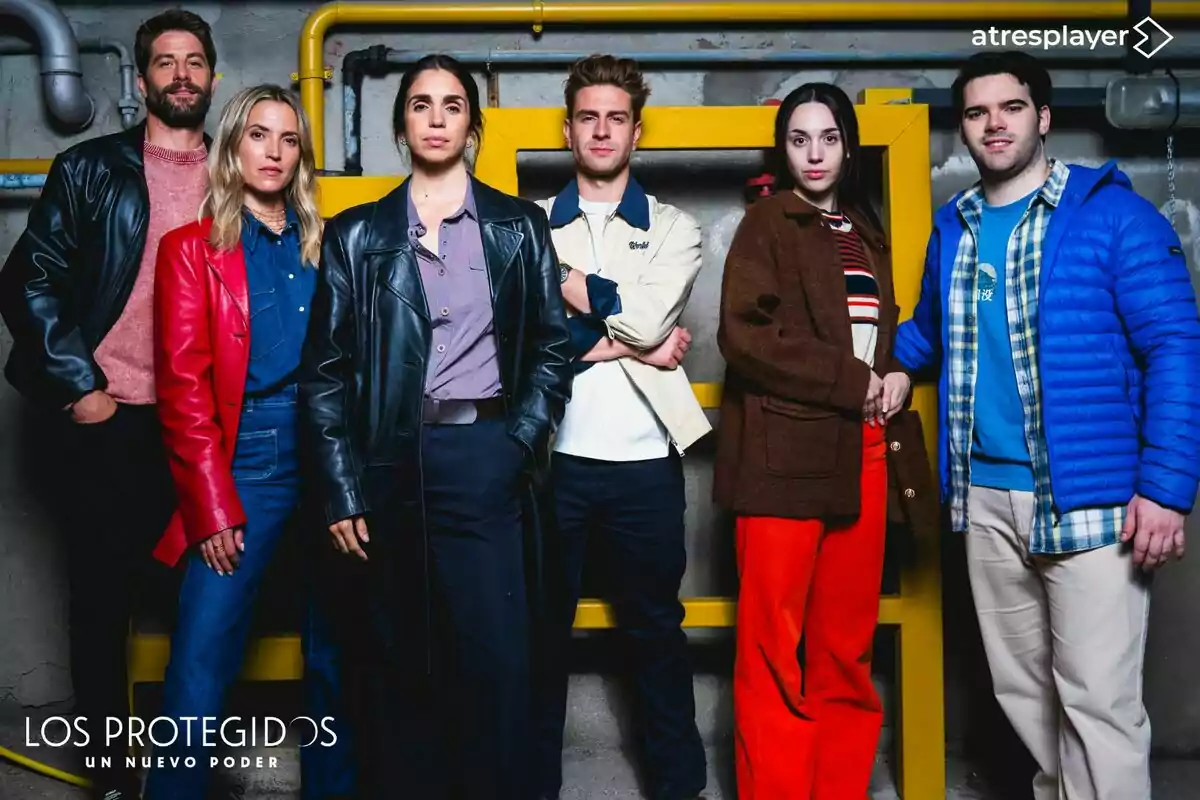 Ana Fernández, Luis Fernández, Mario Marzo, Elena Furiase, Daniel Avilés i Maggie García posen juntes davant d’una paret grisa amb canonades grogues, totes vestides amb roba casual i abrics de colors vius, sota el logotip d’una sèrie anomenada Els Protegits Un Nou Poder i el logotip d’atresplayer a la cantonada superior dreta.