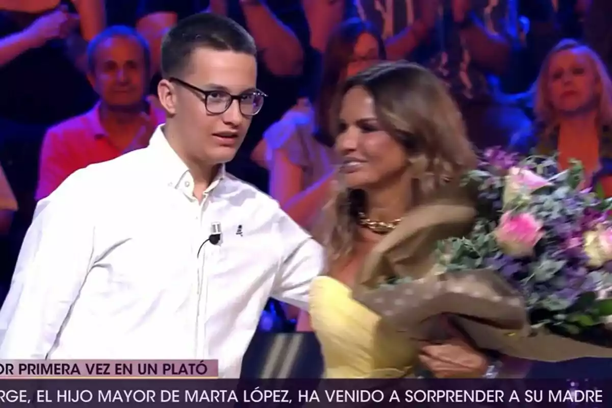 Marta López, que sosté un ram de flors, és abraçada pel seu fill, un jove amb ulleres i camisa blanca, en un plató de De Divendres amb públic al fons. Marta López, que sosté un ram de flors, és abraçada pel seu fill, un jove amb ulleres i camisa blanca, en un plató de De Divendres amb públic al fons.