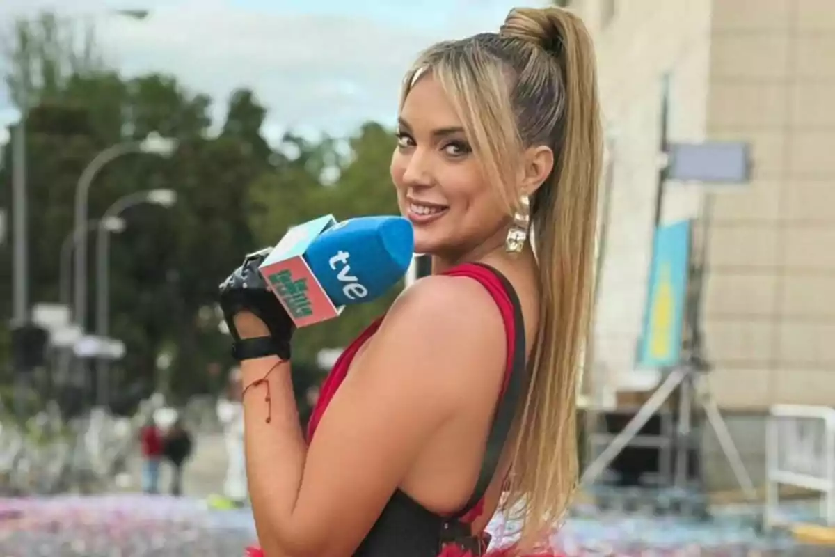 Marta Riesco rossa amb cua alta i vestit vermell sostenint un micròfon blau de tve mentre somriu a la càmera en un esdeveniment a l’aire lliure Marta Riesco rossa amb cua alta i vestit vermell sostenint un micròfon blau de tve mentre somriu a la càmera en un esdeveniment a l’aire lliure