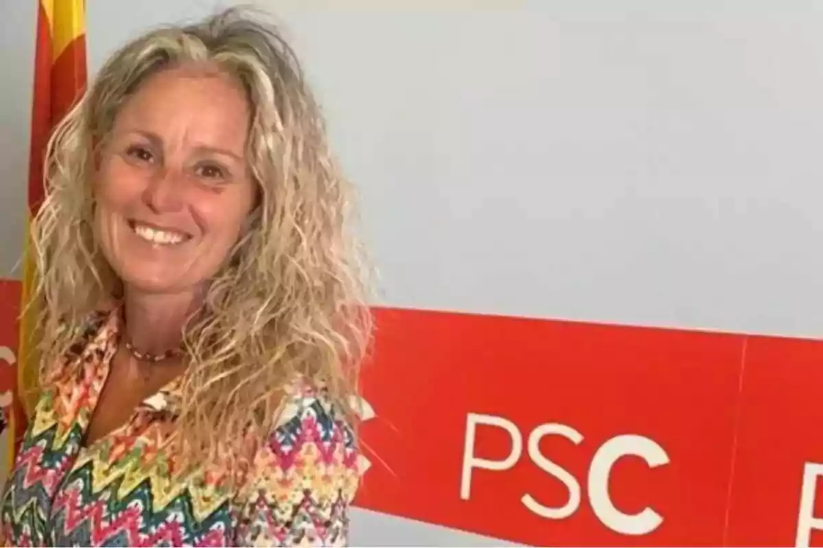 Dona somrient de cabell ros arrissat amb una camisa colorida posant davant d’un fons amb el logotip del PSC i una bandera. Dona somrient de cabell ros arrissat amb una camisa colorida posant davant d’un fons amb el logotip del PSC i una bandera.
