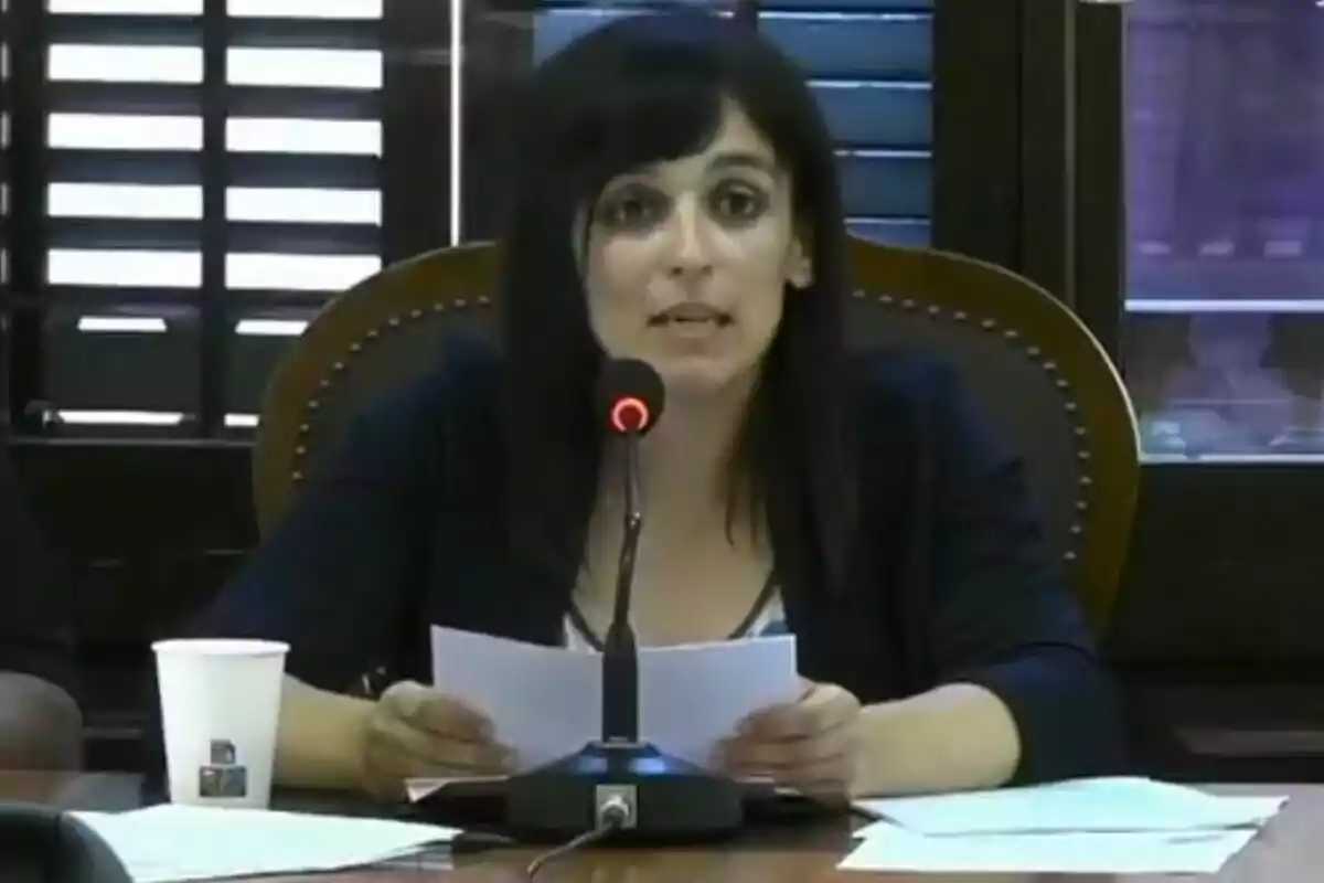 Dona asseguda en una taula parlant davant d’un micròfon amb papers a les mans i un got de cartró al seu costat