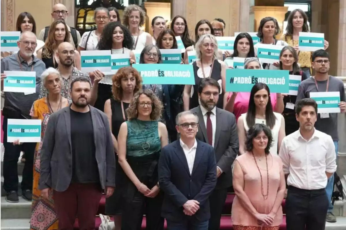 Un grup de persones posant en una escala, moltes d’elles sostenen cartells verds amb el missatge PADRÓ ÉS OBLIGACIÓ Un grup de persones posant en una escala, moltes d’elles sostenen cartells verds amb el missatge PADRÓ ÉS OBLIGACIÓ