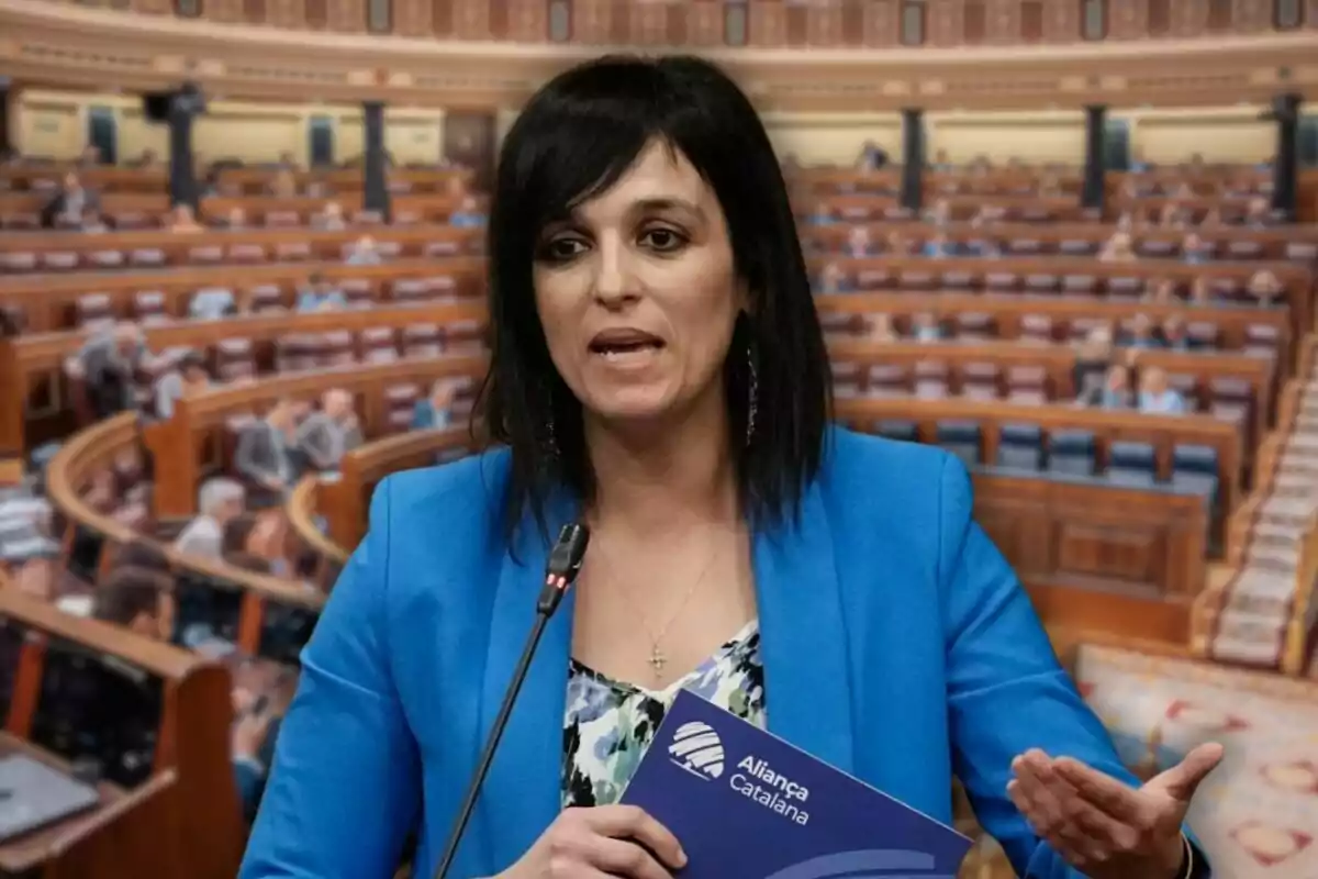 Dona de cabells foscos amb jaqueta blava parlant en un parlament mentre sosté una carpeta d’Aliança Catalana
