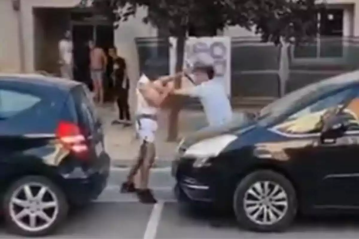 Dues persones discuteixen i s’empenyen entre dos cotxes aparcats al carrer mentre diverses persones observen des de la vorera.