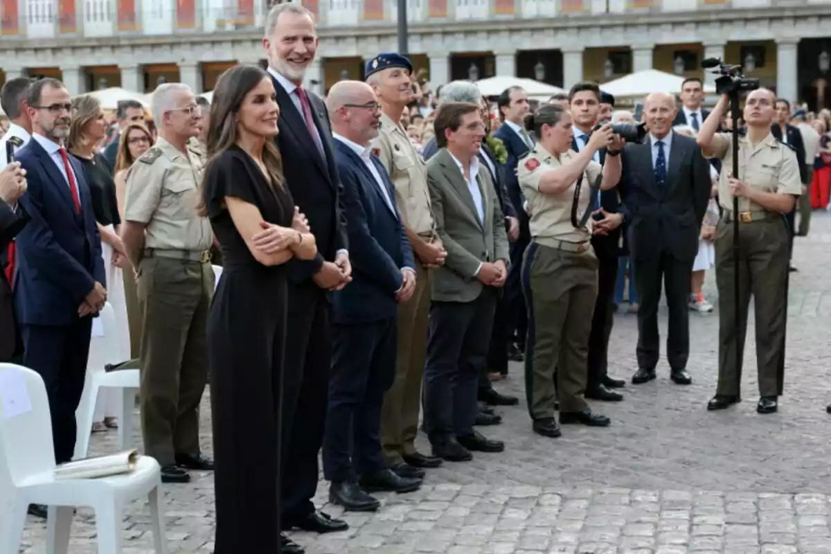 Un grup de persones vestides de manera formal i militar es troba dret en una plaça a l’aire lliure mentre observen i fotografien un esdeveniment. Un grup de persones vestides de manera formal i militar es troba dret en una plaça a l’aire lliure mentre observen i fotografien un esdeveniment.