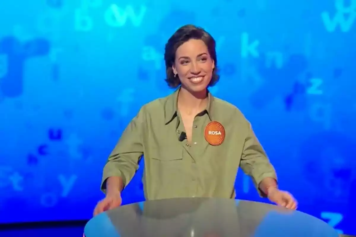 Rosa Rodríguez, smiling, wearing a green jersey and an orange name tag that says Rosa, participating in a television show called 'Pasapalabra', with a blue background full of letters. Rosa Rodríguez, smiling, wearing a green jersey and an orange name tag that says Rosa, participating in a television show called 'Pasapalabra', with a blue background full of letters.