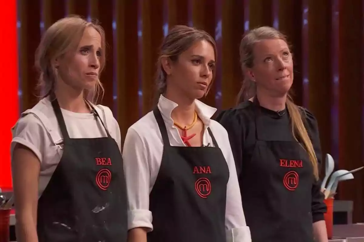 Tres dones amb davantals negres de MasterChef i noms brodats en vermell estan dretes en una cuina amb expressions serioses i concentrades
