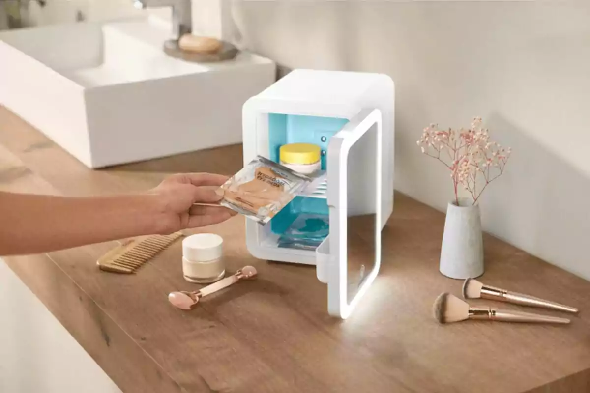 Una persona guarda productos de cuidado facial en un mini refrigerador sobre una mesa de madera junto a brochas de maquillaje, un peine y un jarrón pequeño con flores secas. Una persona guarda productos de cuidado facial en un mini refrigerador sobre una mesa de madera junto a brochas de maquillaje, un peine y un jarrón pequeño con flores secas.