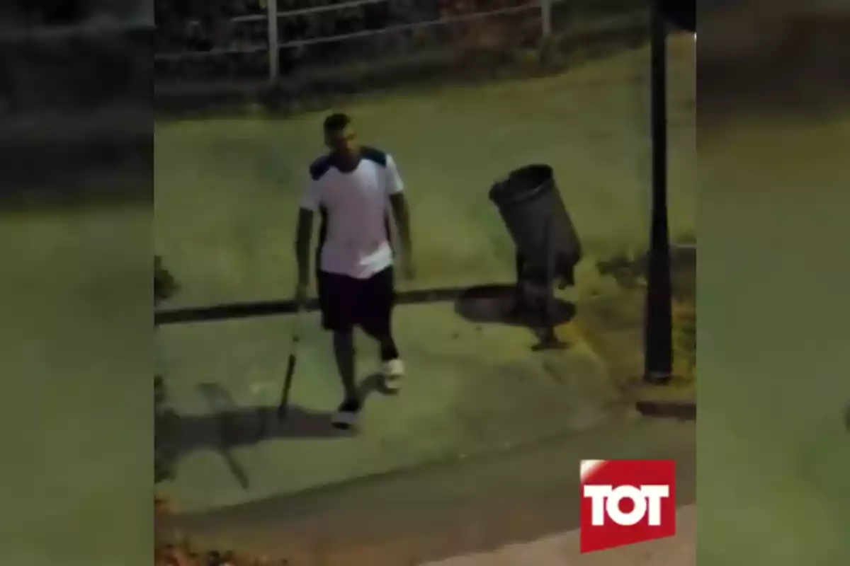 A man in a white t-jersey and dark shorts walks on the sidewalk at night holding a long object near a trash can and a light pole.