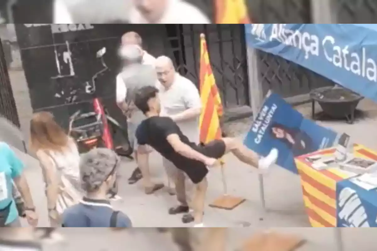 A young man kicks a political propaganda sign next to a table decorated with the Catalan flag on a busy street.