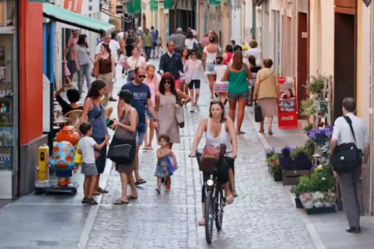 Busy pedestrian street with people walking and a woman riding a bicycle among shops and flowers
