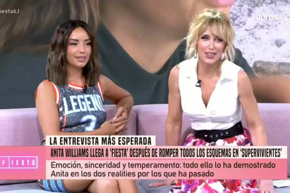 Two women sit on a sofa during a Fiesta show, Anita Williams wearing a sleeveless dress with the word LEGEND and Emma García in a white blouse and colorful skirt, both smiling as text appears on the screen about an anticipated interview.
