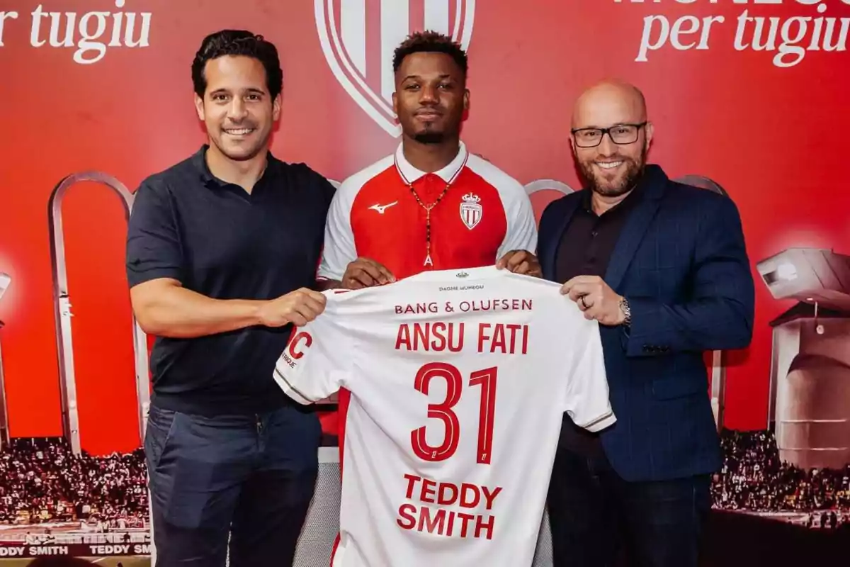 Tres persones posen somrients mentre sostenen una samarreta de futbol del Mònaco amb el nom d’Ansu Fati i el número 31 davant d’un fons vermell amb l’escut del club. Tres persones posen somrients mentre sostenen una samarreta de futbol del Mònaco amb el nom d’Ansu Fati i el número 31 davant d’un fons vermell amb l’escut del club.