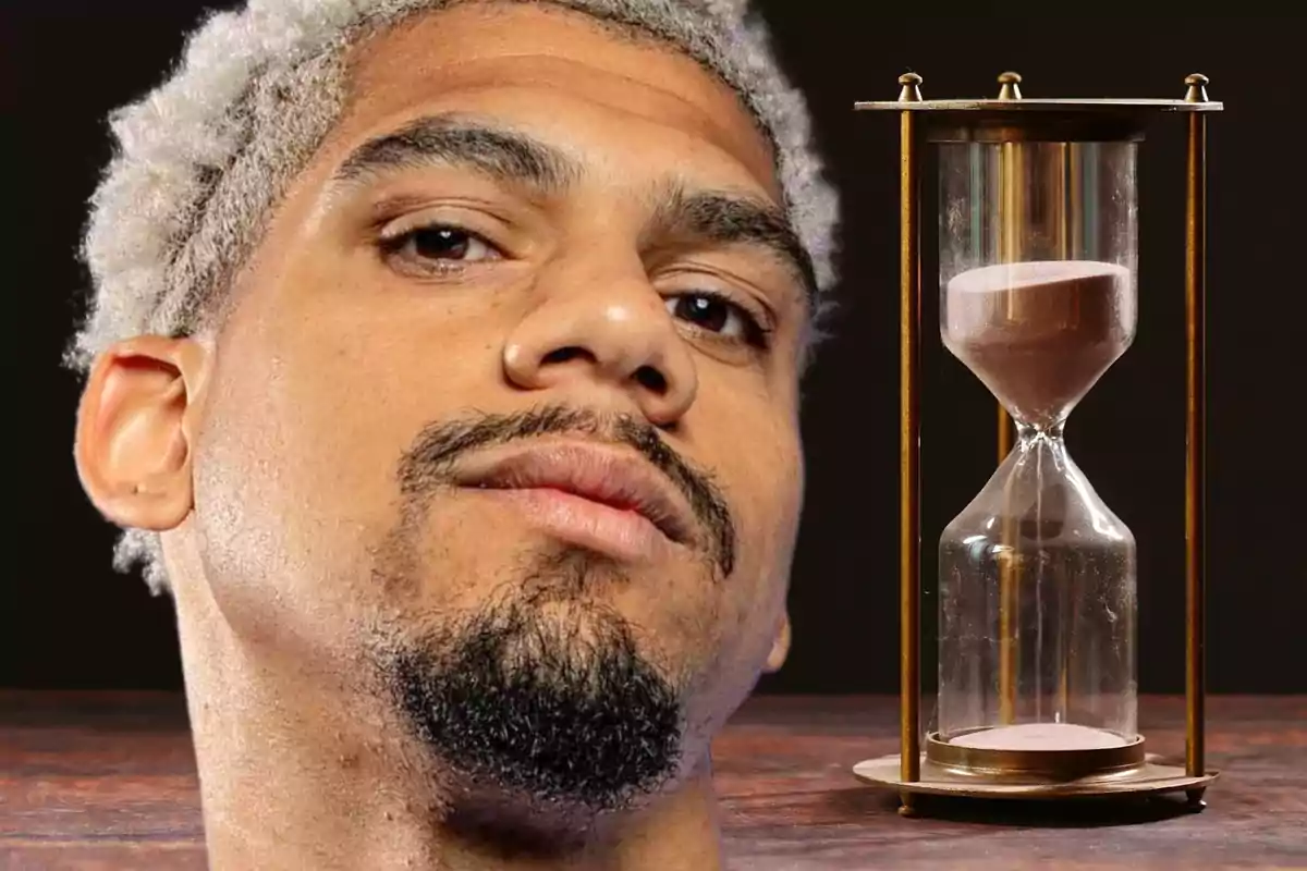 A curly-haired man with a beard looks at the camera next to an hourglass on a wooden table.