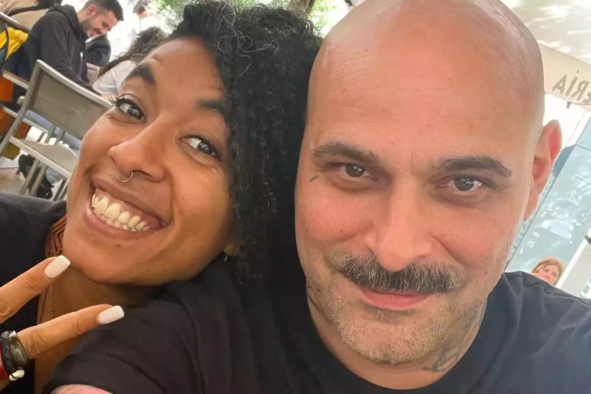 Dues persones somriuen i posen juntes per a una selfie en un lloc a l’aire lliure, una d’elles fa el signe de la pau amb els dits.